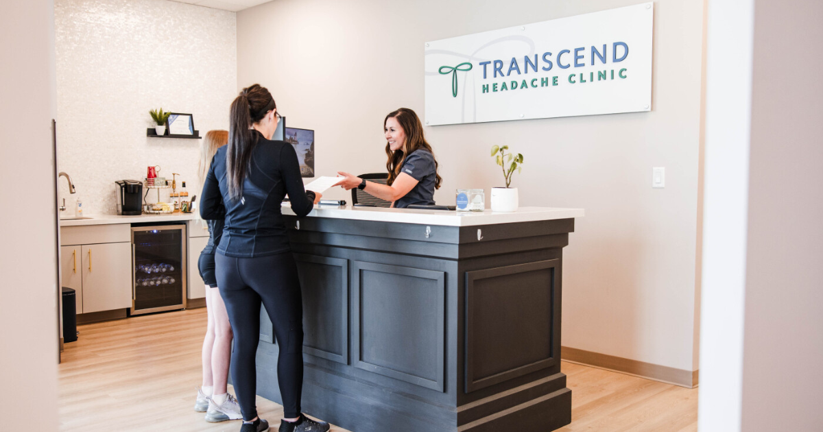 A new patient checks in at the front desk of Transcend Headache Clinic in West Fargo, ND, as a smiling staff member hands over paperwork beneath the clinic's sign.