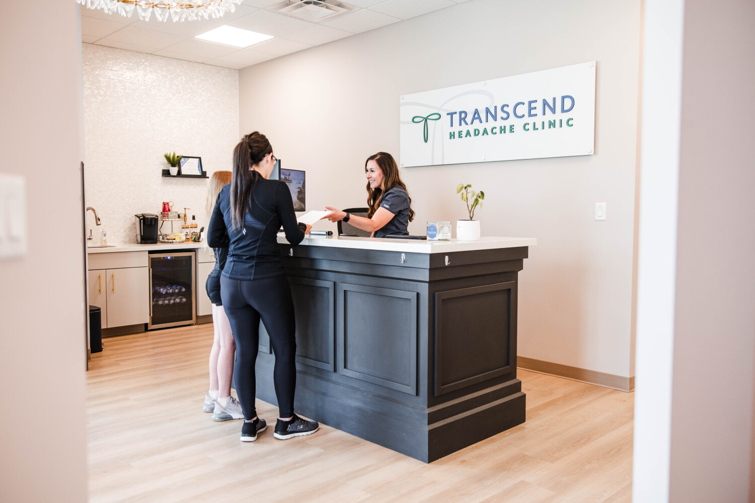 A new patient checks in at the front desk of Transcend Headache Clinic in West Fargo, ND, as a smiling staff member hands over paperwork beneath the clinic's sign.